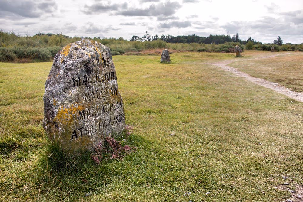 A clan memorial stone standing in the grass on Culloden moor, the path and the plantation behind it