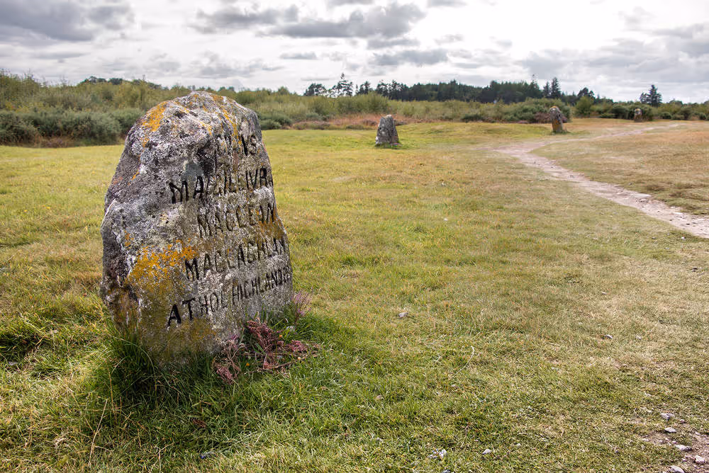 A clan memorial stone standing in the grass on Culloden moor, the path and the plantation behind it