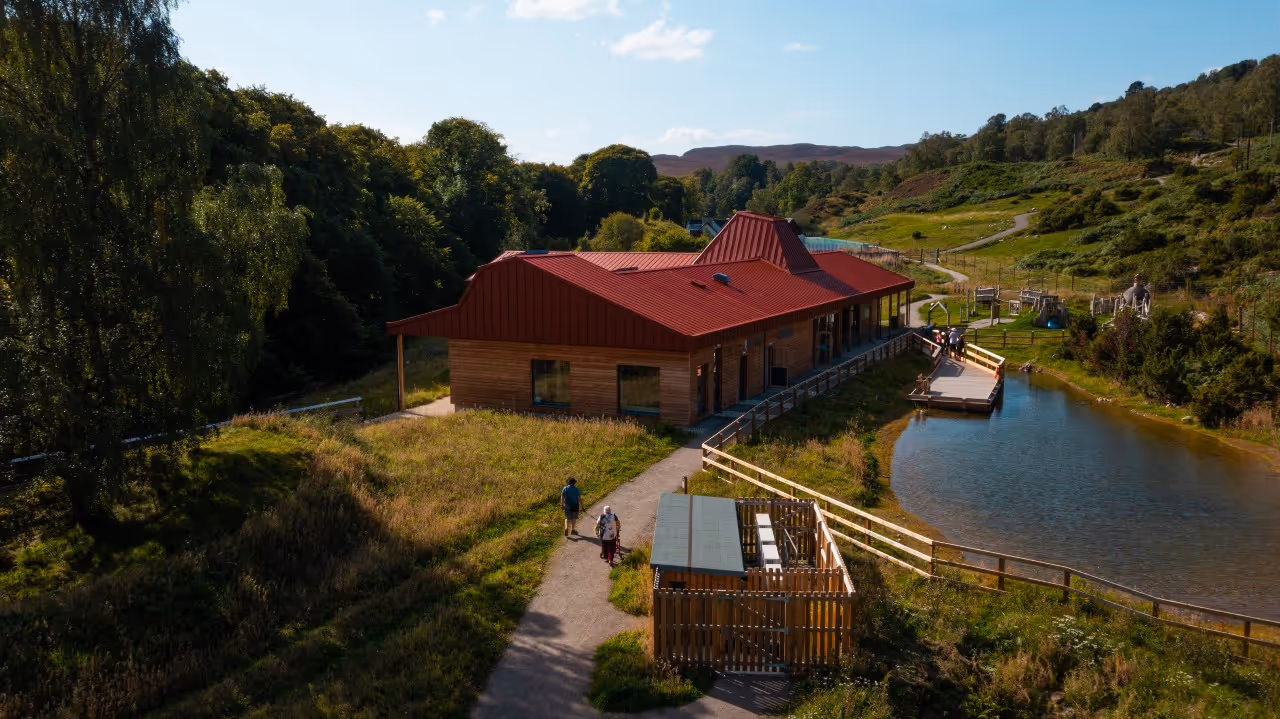 The rewilding centre at Dundreggan, a low timber building with a red roof above a pond and boardwalk, paths running out into the birch and open hill behind