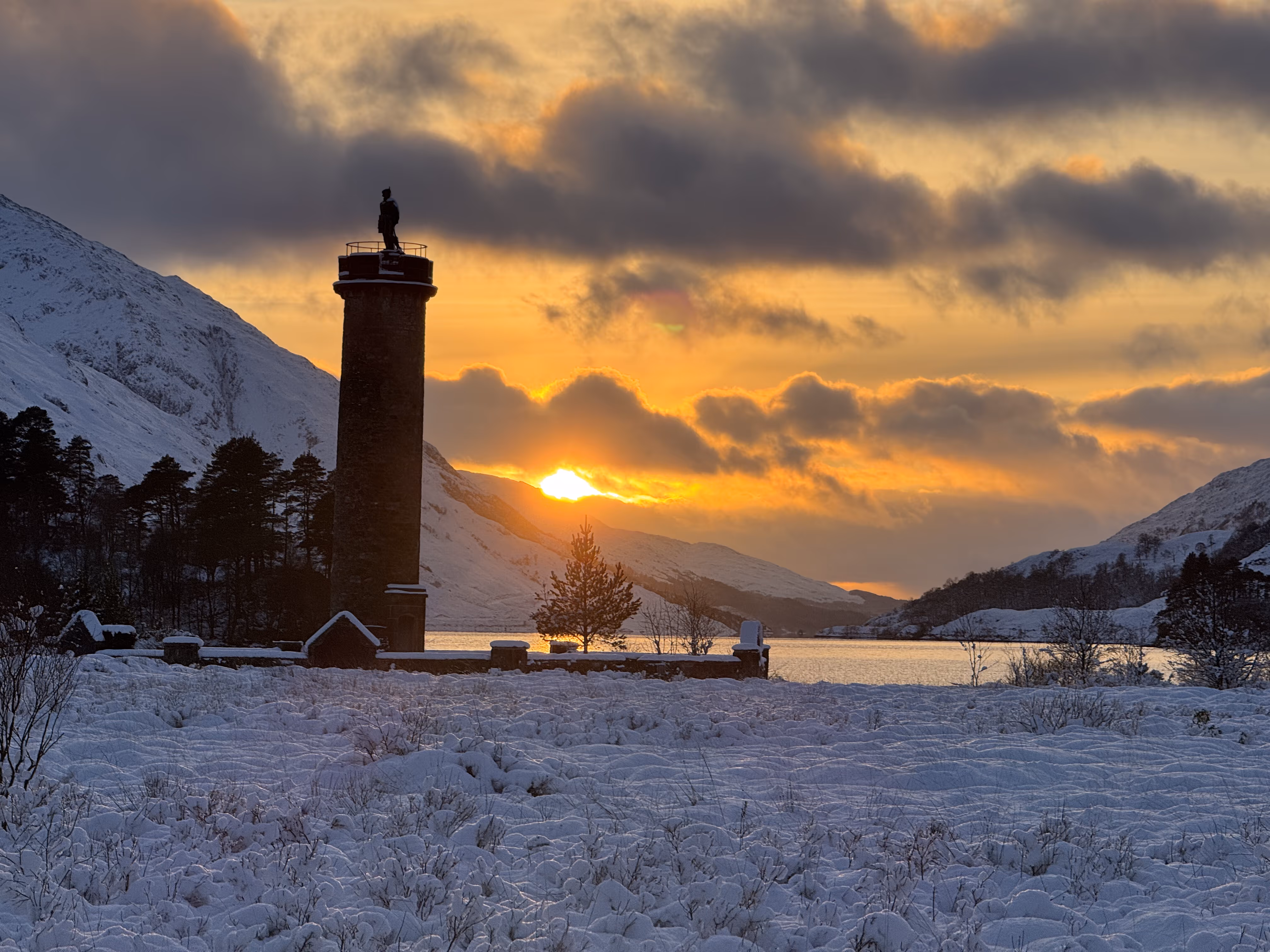The Glenfinnan monument in silhouette against a January sunset over Loch Shiel, snow on the ground and along the hills on both sides