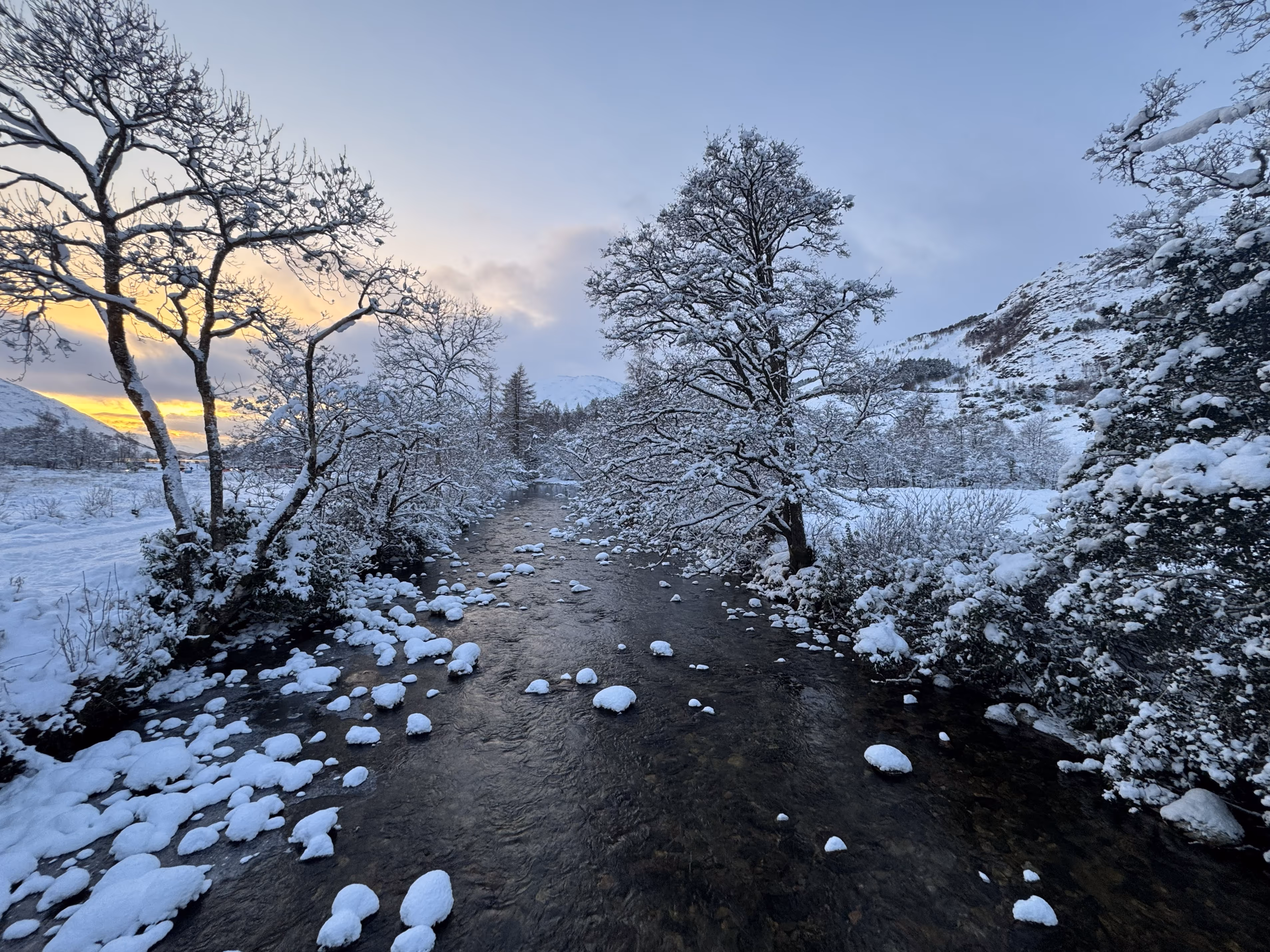 The river at Glenfinnan under snow at dusk, snow-laden birches leaning over the water and boulders capped white in the shallows