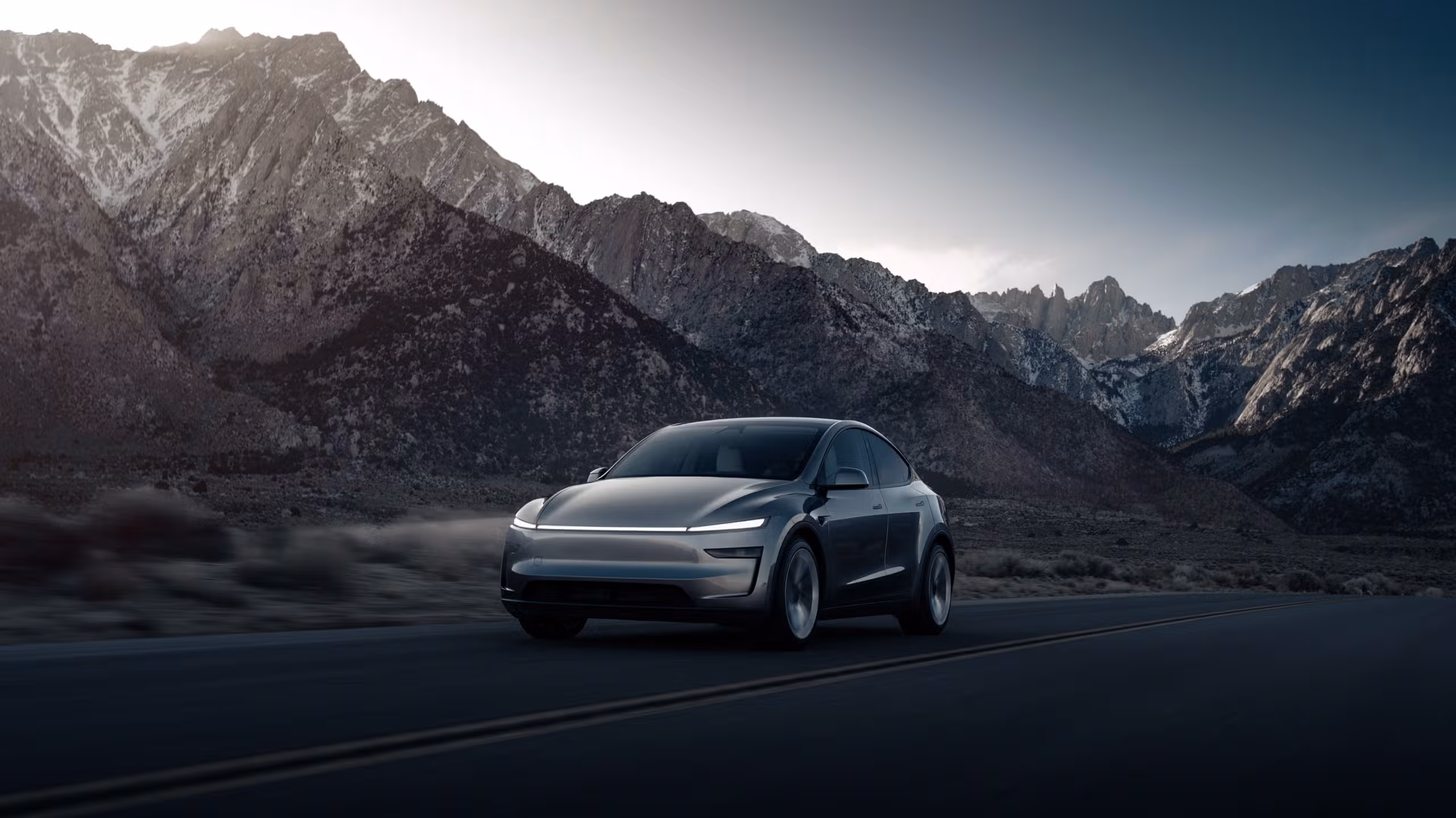A Tesla Model Y on an empty mountain road at dusk, jagged peaks rising behind it
