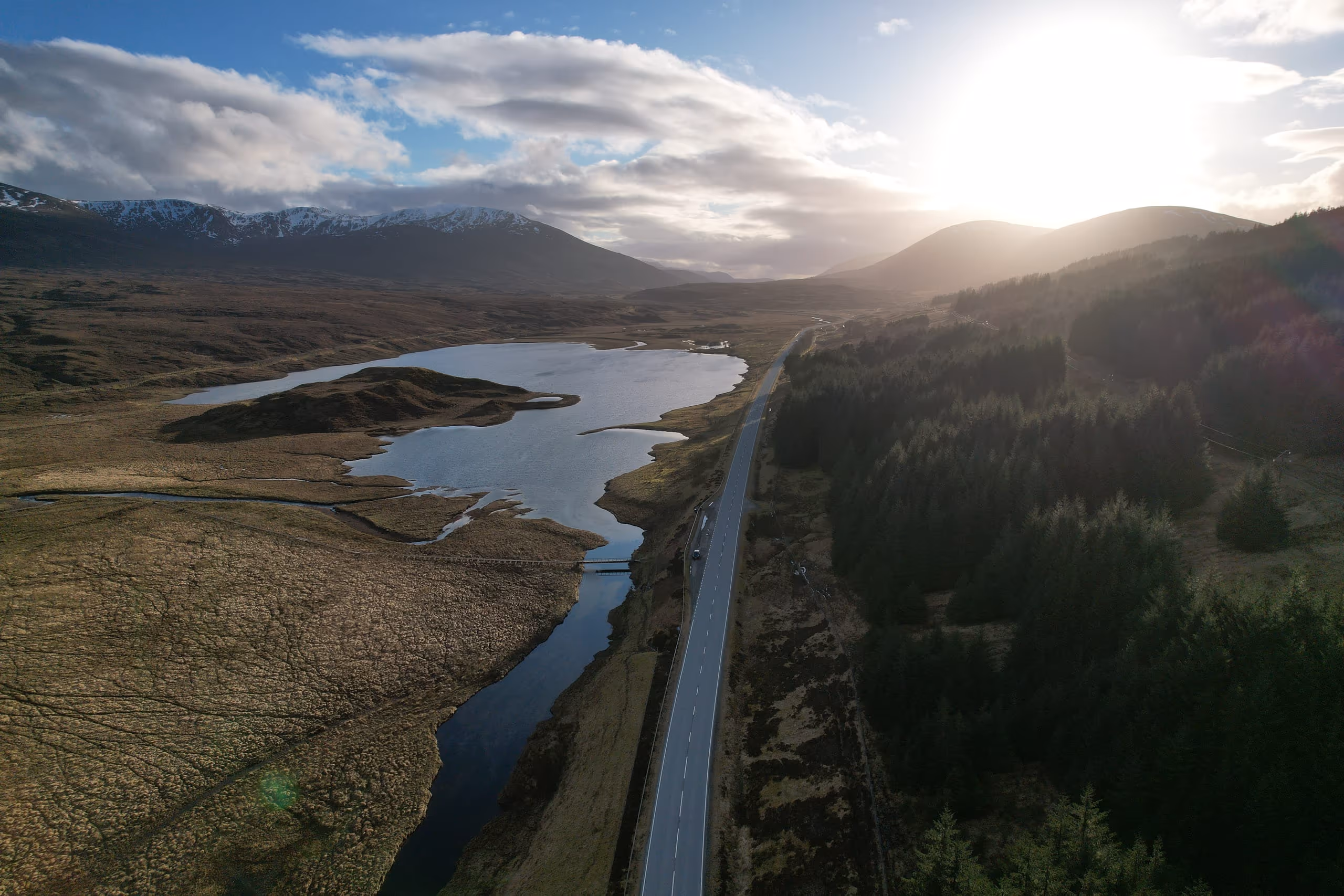 A single-track Highland road running away through a glen beside a loch, low sun over the hills at the end of it