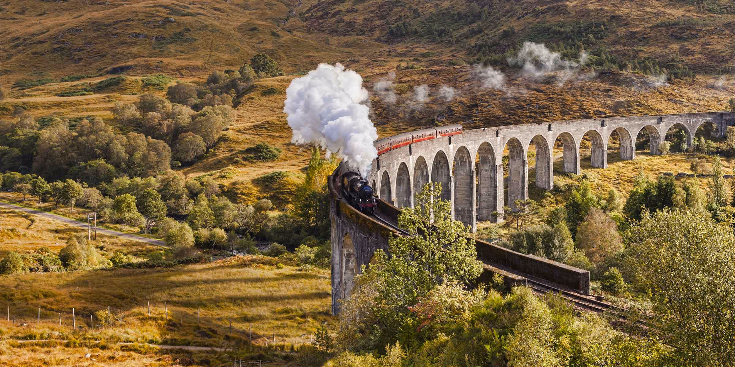 The Jacobite steam train crossing the Glenfinnan viaduct in autumn, steam rising against the hillside