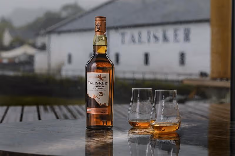 A bottle of Talisker and two filled glasses on a table, the white distillery buildings at Carbost behind