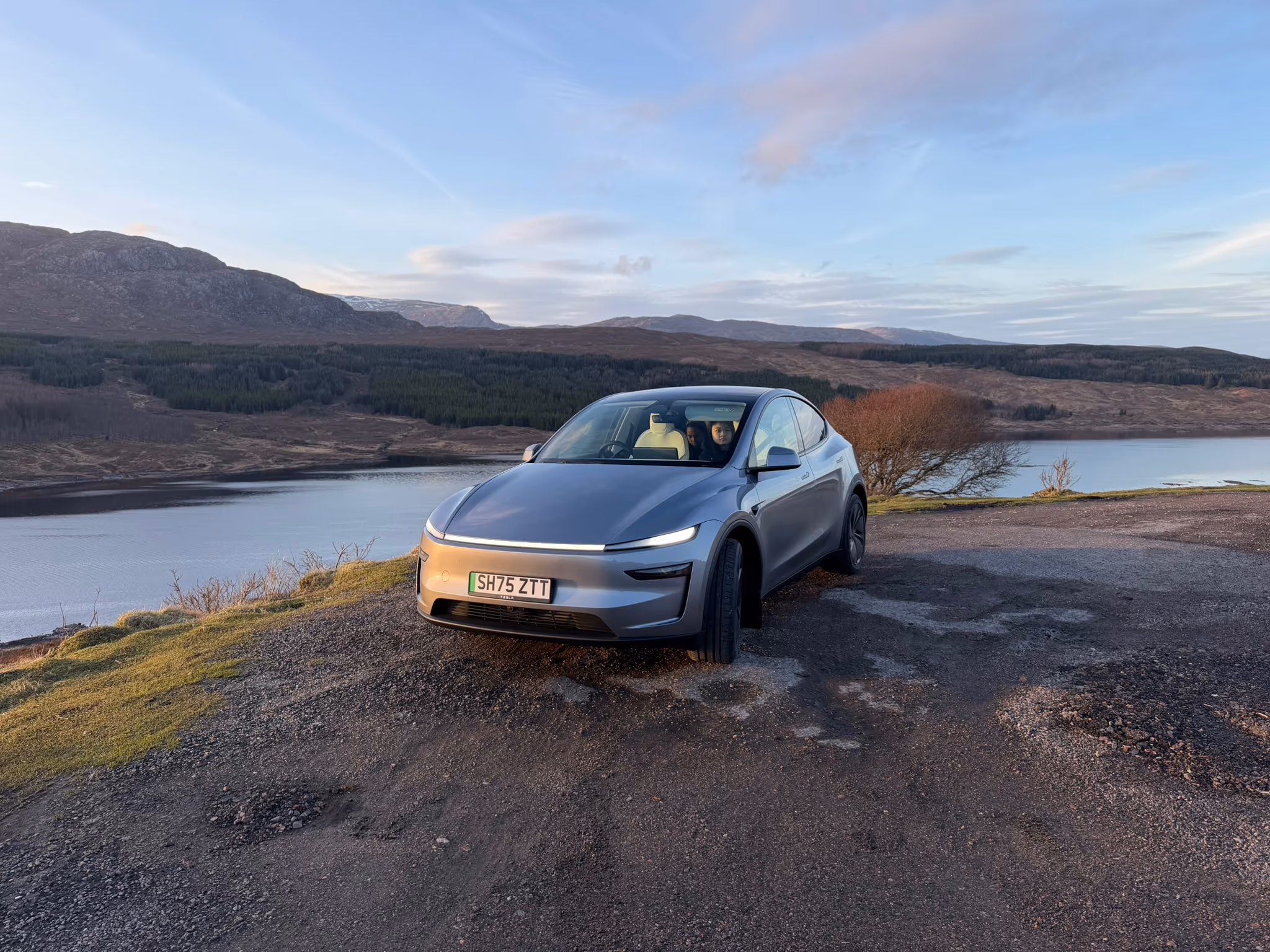 The silver Tesla parked at the Loch Loyne viewpoint on a still winter afternoon, the loch low below and snow on the tops behind